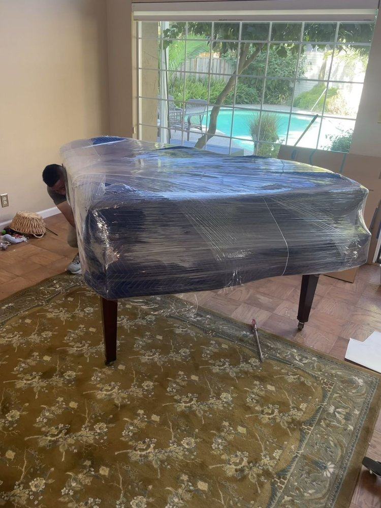 A mover preparing a plastic-wrapped grand piano inside a home
